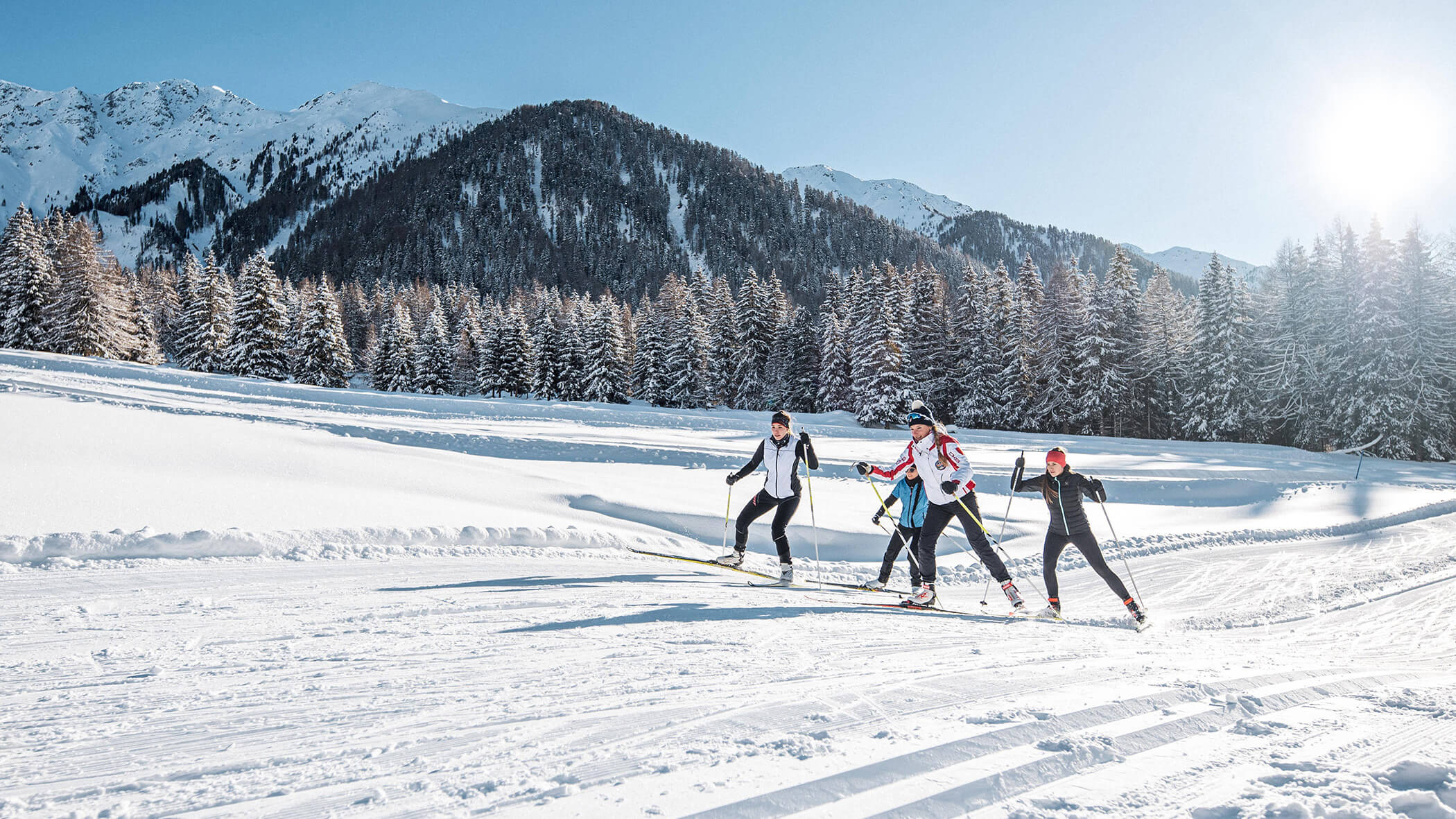 Vier Langläufer auf der Loipe, im Hintergrund schneebedeckte Berge und Bäume - Oberplunerhof