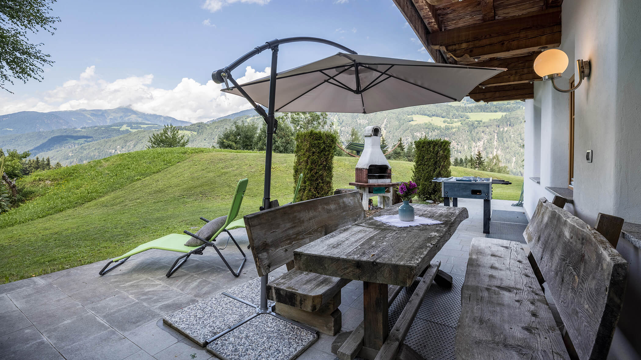La terrazza dell'Oberplunerhof con tavolo e panche in legno, barbecue e sedia a sdraio sullo sfondo